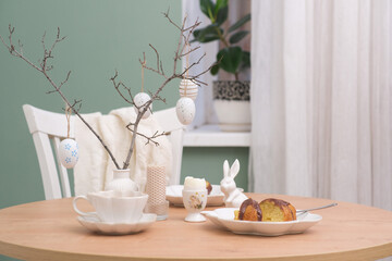 Easter table. Vase with fresh branches, cup of tea, cupcake on plate, boiled egg, ceramic bunny, candle on wooden table with white chair on curtain background.