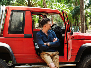 Attractive man and a red SUV against a background of palm trees. Sunny, clear day. Closeup, outdoor. Vacation and travel concept © Svetlana