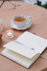 opened notebook with white pen, a cup of tea and burning candle on wooden table and chair. Top view.