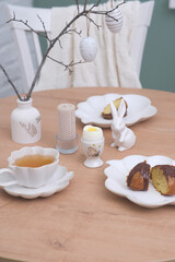 Easter table. Vase with fresh branches, cup of tea, cupcake on plate, boiled egg, ceramic bunny, candle on wooden table with white chair on curtain background.