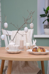 Easter table. Vase with fresh branches, cup of tea, cupcake on plate, boiled egg, ceramic bunny, candle on wooden table with white chair on curtain background.