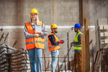 Portrait of successful woman constructor wearing yellow helmet and safety yellow vest. Portrait of architect standing at building site and looking at camera.