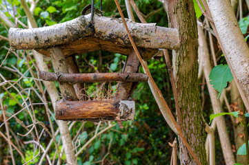 A wooden birdhouse made of birch branches hanging from the branches of a bush.