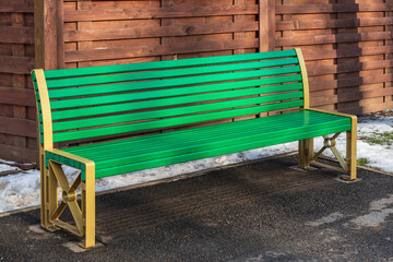 Modern green bench in a park on the asphalt near a wooden fence