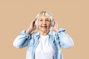 Cheerful elderly blonde woman lady 60s years old in casual clothes listening music with headphones isolated on pastel beige color background, Energetic senior pensioner