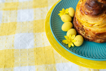 A delightful pastry on a teal plate with yellow Easter eggs and a flower, set on a checkered tablecloth.
