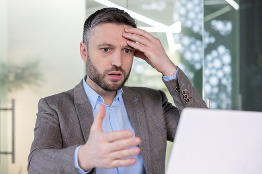A professional businessman appears confused and stressed while working at his laptop in a modern office environment.