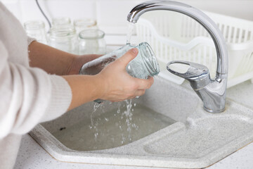 Well in the kitchen, a woman washes glass jars in the sink to sort them for recycling. The woman is...
