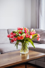 Obraz na płótnie Canvas Vase of fresh tulips on the coffee table with blurred background of modern cozy light living room with gray couch sofa and graphic cushions. Open space home interior design. Copy space. Vertical card.
