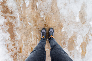 Legs in denim jeans encased in grey shoes stand on snowcovered ground