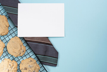 Necktie and blank card lying beside of fresh baked chocolate chip cookies. Light blue background.