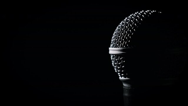 The vocal handheld microphone slowly rotates in backlight on black background. Close-up chrome grid on dynamic mic surface. Copyspace. Concept recording studio, voice, podcast, karaoke, audiobook