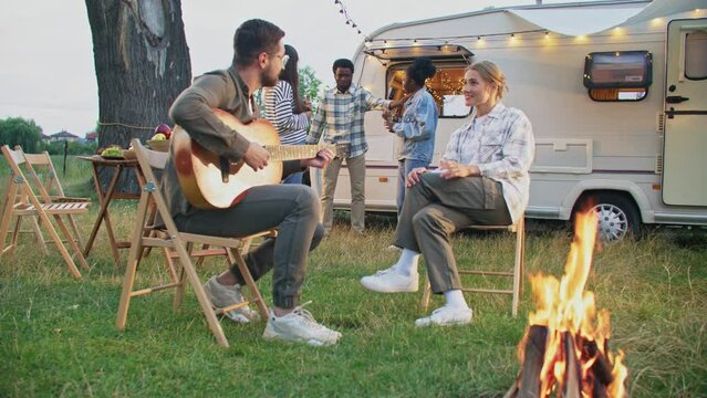 Mixed-raced people having party in nature. Handsome man playing on guitar romantic sound while woman listening. Loving couple sitting near flame. Friends communicating next to trailer in background.