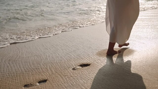 Barefoot person in flowing dress walks along sandy shoreline. Waves gently wash over footprints. Peaceful beach stroll at sunset provides tranquil moment, reflection. Slow motion.