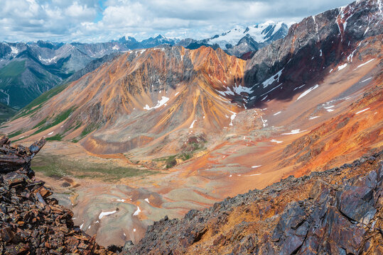 Awesome scenic top view from precipice edge with sharp rocks to unusual multicolor stony valley with iron river among colorful high mountains and large snowy mountain range in sunlight in cloudy sky.