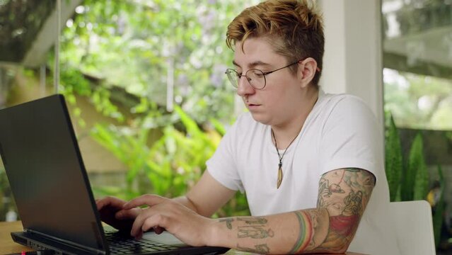 Tattooed transgender professional types on laptop at plant-filled coworking space. Focused individual with glasses engages in remote work, symbolizing inclusivity and modern freelancing. Slow motion.