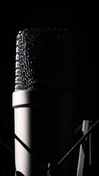 Vertical, studio microphone rotates on a black background in backlight close-up. Condenser microphone with chrome grid on surface. Concept recording studio, voice, podcast, karaoke. Copy space