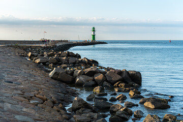 Large breakwaters at the Warnow estuary. The pier fire on the west pier in the background. Evening atmosphere on the promenade in Warnemünde in Mecklenburg-Western Pomerania. Walkers on the pier. - obrazy, fototapety, plakaty