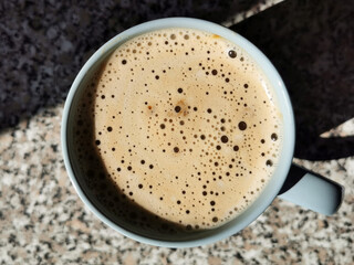 Top view of a blue coffee mug with beige milk foam with bubbles of different sizes, which stands on a mottled chipboard countertop with a stone texture