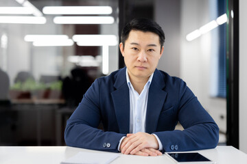 Portrait of a confident Asian businessman seated at a table in a well-lit, contemporary office...