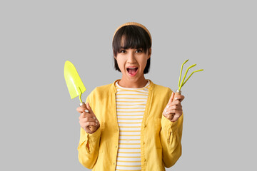 Beautiful young shocked female gardener with gardening rake and shovel on grey background