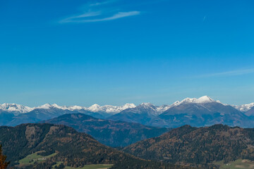 Fototapeta na wymiar Panoramic view of snow capped mountain ridges of Woelzer Tauern seen from Grebenzen, Gurktal Alps, Styria, Austria. Calm serene atmosphere in Austrian Alps. Idyllic forest in foreground. Wanderlust