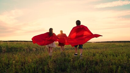 Happy girls, boy kids play superheroes they run across green field in red cloak, cloak flutters in...