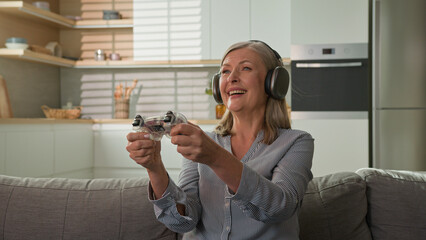 Funny smiling mature caucasian woman playing video game console in headphones using joystick controller at home on couch elderly female play computer games older people and modern technology concept - obrazy, fototapety, plakaty