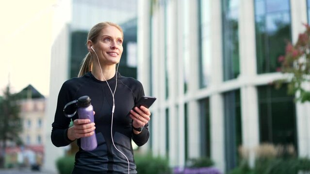 Happy adult sportswoman using a phone walking in city street. Fit female runner with headphones enjoying listening to music outdoors. Joyful athlete in black sportswear returning from training or run