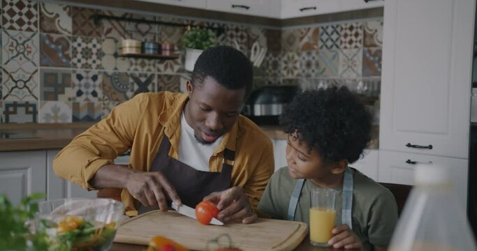 Happy family dad and son making delicious salad cutting tomato sitting at table in kitchen talking laughing. Nutrition and fatherhood concept.