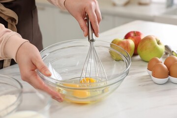 Woman whisking eggs in bowl at light marble table indoors, closeup