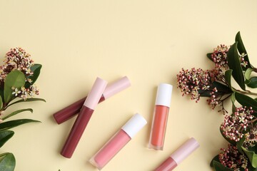 Different lip glosses and flowers on pale yellow background, flat lay