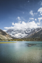 Naklejka na ściany i meble Blue mountain lake in a mountain valley among vegetation against the backdrop of rocky peaks with snow in the Fan Mountains in Tajikistan, Tien Shan highlands in the evening
