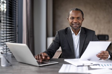 Professional African American businessman analyzing paperwork with laptop and tablet on his desk in...