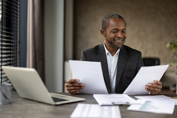 A professional in a suit carefully analyzes documents, surrounded by a laptop and reports on a desk in a well-lit office.