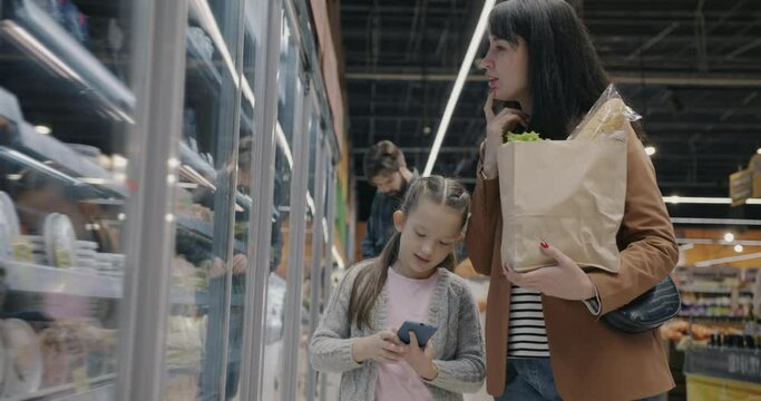 Young family mum and child shopping for food holding paper bag talking and using smartphone in supermarket. People and consumerism concept.