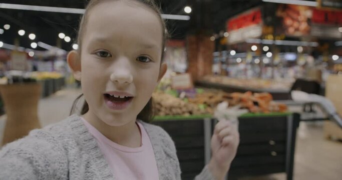 Portrait of adorable child making online video call from supermarket talking waving hand looking at camera showing food. Childhood and internet communication concept.