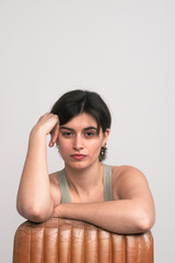 A young woman casually supports her head with her hand while leaning against a brown leather chair, gazing directly at the camera.