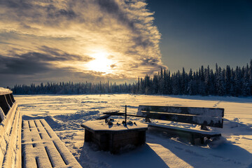 Une table brasero et deux bancs devant une forêt et une étendue de neige au lever du soleil en Laponie en Suède - obrazy, fototapety, plakaty