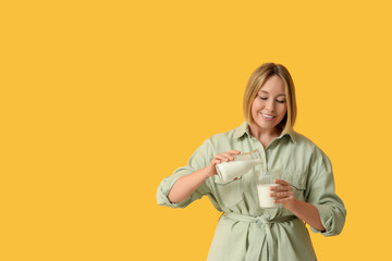 Beautiful young woman pouring fresh milk from bottle into glass on yellow background