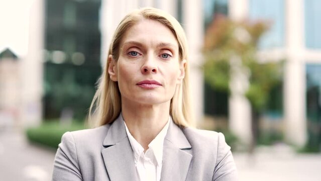 Portrait of a serious businesswoman standing on street near an office building. Adult blonde woman in a formal suit posing looking at camera. Headshot of confident mature female manager. Close up