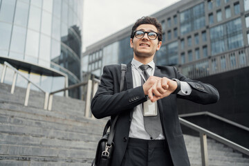 A hurried businessman checks his watch against the backdrop of a modern office complex, embodying...