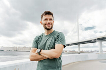 Fototapeta premium A contented man with crossed arms gazes into the distance, a smartwatch on his wrist, set against an expansive bridge and cloudy sky backdrop.