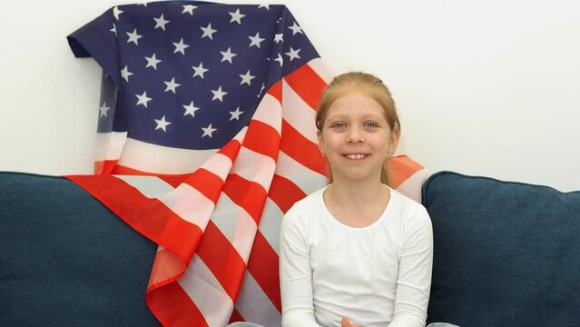 The girl speaks to the camera. US flag in the background. Storytelling about American education. Studying of foreign language. Child with American flag. Independence Day and US National Flag Day