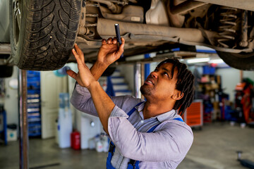 African-American mechanic beneath a car on a lift, using a handheld lamp to illuminate and identify...