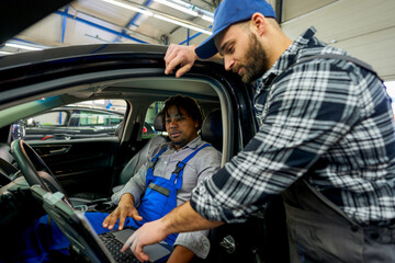 Mechanic colleagues connected the car to diagnostics. One in the driver's seat, the other outside, discussing findings on the computer screen, working together to identify the issue.