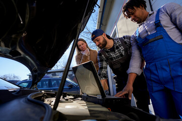 A worried woman, flanked by mechanic colleagues, scrutinizes a car using computer diagnostics. They...