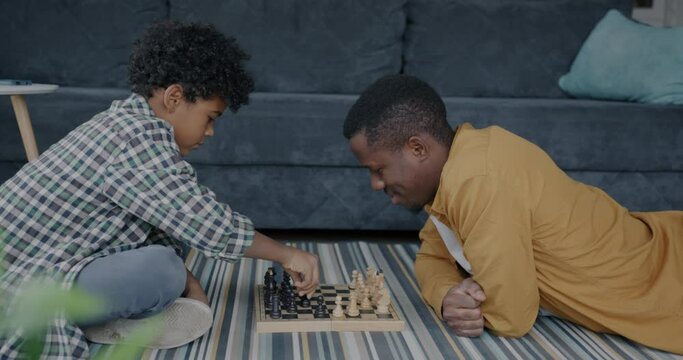 Cute child playing chess with caring father and watching adorable kitten on floor in apartment. Family lifestyle and happy childhood concept.
