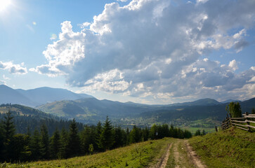 A well-trodden path leads into a valley of the Verkhovyna village nestled amidst rolling hills, adorned with lush greenery under the sky is painted with clouds filtering the golden sunlight