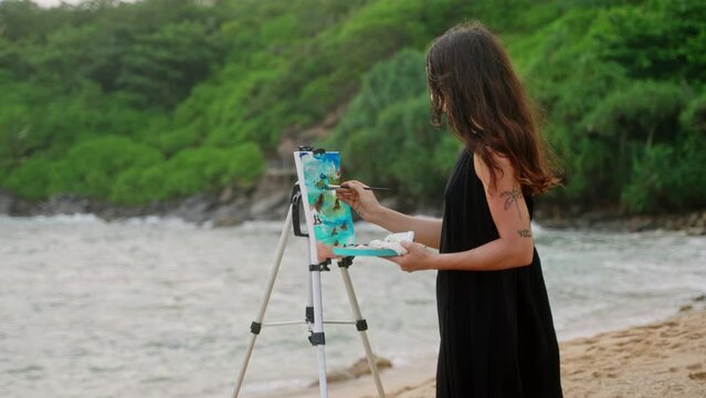 Woman paints seascape on canvas at tropical beach. Artist creates artwork with brush, color palette, ocean waves backdrop. Art inspiration in natural outdoor setting. Creative hobby, travel theme.